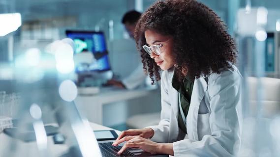 woman in white medical coat typing on a laptop in lab setting