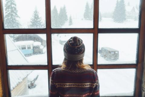 Person in sweater and knit hat facing window, with winter, snowy yard outside