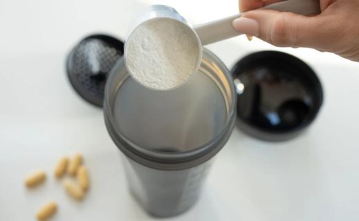 Hand pouring scoop of supplement powder into shaker, with blurry pills on the counter