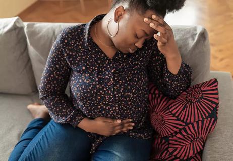 A pregnant person sits on the couch holding their stomach and head with irritation.