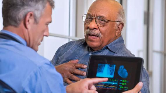 Older male in doctor's office with doctor holding tablet showing heart statistics