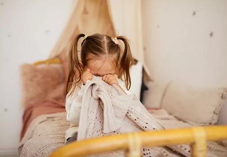 Toddler sitting on bed, hiding her face behind a blanket.