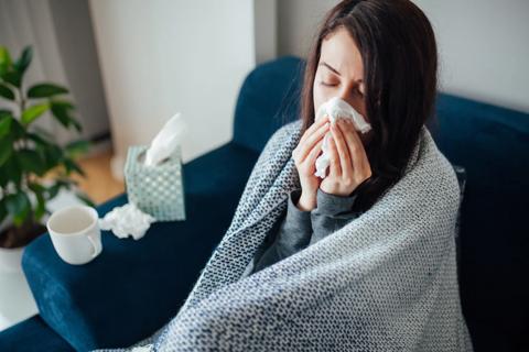 Person wrapped in blanket on couch, blowing their nose, box of tissues and mug of warm drink nearby