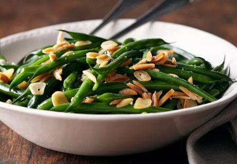 Green beans topped with nuts in a white bowl displayed on a dark wooden table.