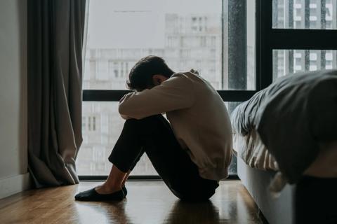 Person sitting on floor in bedroom, dimly lit from the window, with head resting on knees