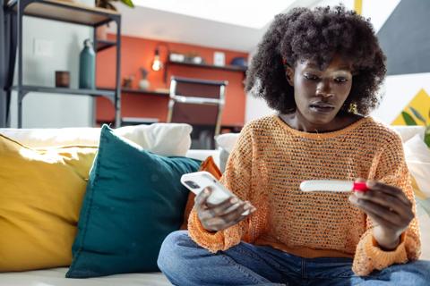 Female sitting on couch looking at a pregnancy test stick, holding cell phone
