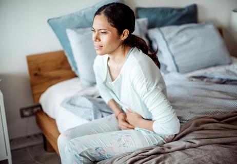 Woman in pajamas sitting on side of bed suffering from abdominal pain.