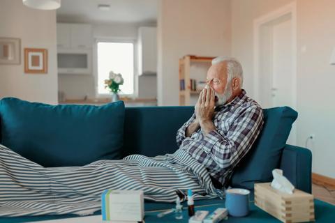 Older male reclining on couch, blowing nose, with cold medications and products on coffee table