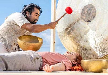 Music practitioner lightly hits gong as patient lies relaxed on matt while sound waves move around body.