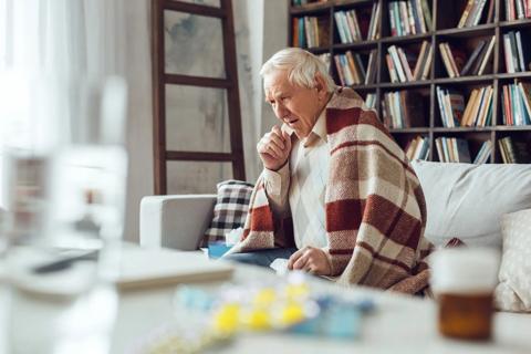 Older person wrapped in blanket sitting on couch, coughing, sick