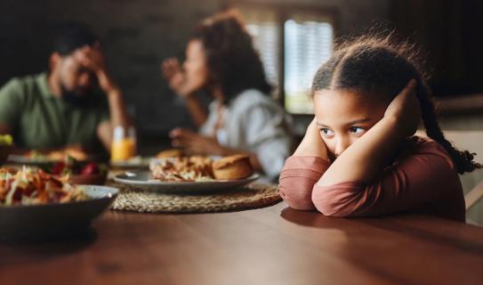 Child covering their ears at dinner table as parents argue next to them