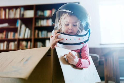 Young child at home, with space helmet on, sitting in cardboard box, coloring on the box