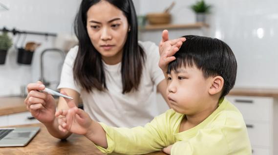 Caretaker reading a digital thermometer, while placing their hand on a sick-looking child's forehead