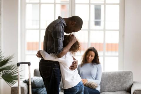 Caregiver standing with suitcase nearby, hugging child, with another caregiver sitting on couch