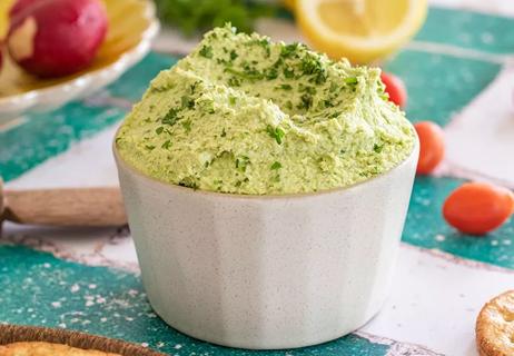 Heaping helping of green hummus flecked with herbs in a white ramekin on a cluttered counter