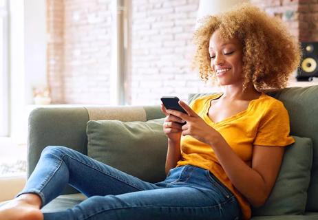Person in yellow tshirt and blue jeans relaxing on green couch in living room reading texts on their phone.