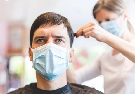 A short-haired man wearing a surgical mask is staring straight ahead while receiving a haircut