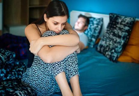 woman upset sitting on bed with man in background