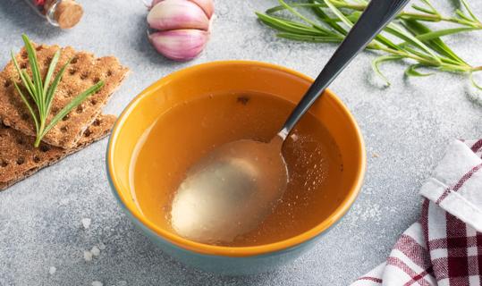 Bowl of broth with spoon, with crackers and herbs on table