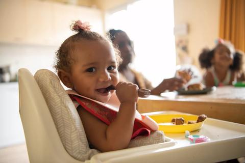 Smiling older baby in high chair with baby utensil in mouth, eating with family