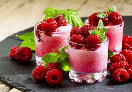 Raspberry smoothies in half glasses topped with fresh raspberries placed on a dark grey tray.