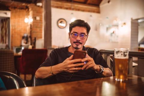 Person sitting at a table in a bar, on their phone, with glass of beer on the table