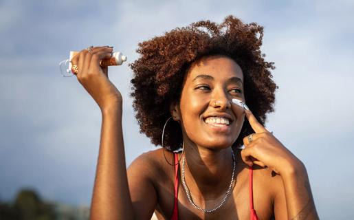 Smiling person under sunny blue sky, holding tube of sunscreen, applying to face