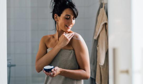 Woman in towel in bathroom after shower, applying moisturizer