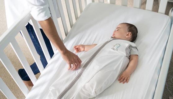 Child in sleeping sack in asleep in crib, with caregiver leaning over, checking on baby