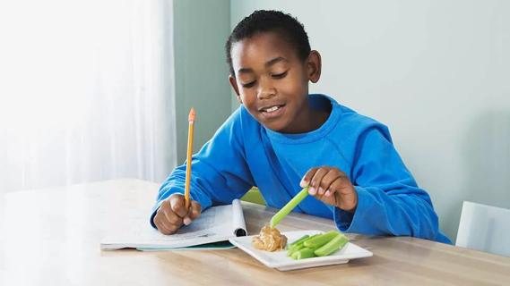 male kid eating celery sticks with peanutbutter at table with homework
