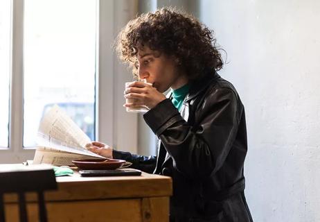 Nonbinary person drinking coffee and reading paper in a coffeeshop.