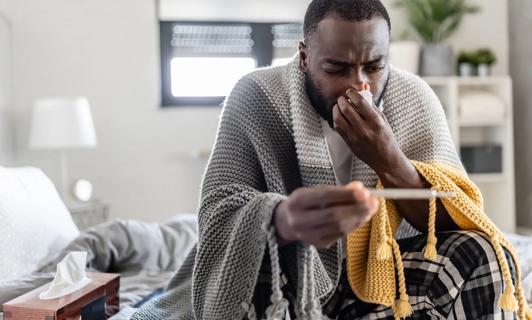 Person home sick, wiping nose with tissue, checking their temp on a thermometer