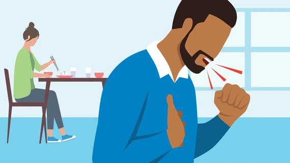 Man steps away from eating at the table to cough with hand on sternum, with partner continuing eating at the table