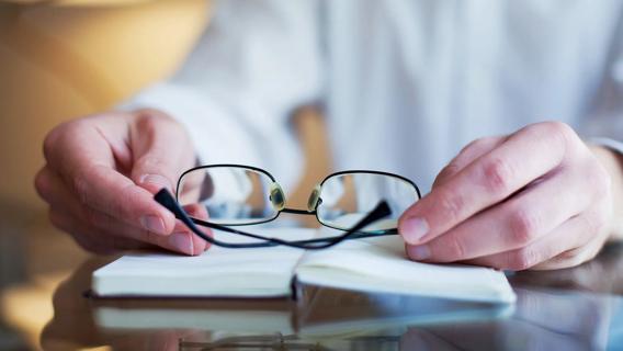 Hands holding "cheater" reading glasses, with a book open on table