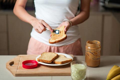 Person spreading peanut butter on soft bread, with glass of milk and bananas nearby