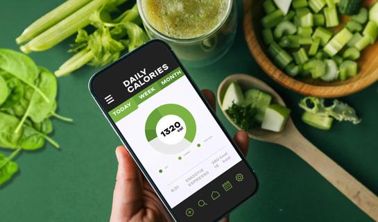 Hand holding a smartphone with a calorie tracking app on it, with greens on a table