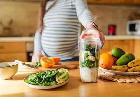 A pregnant person using a blender with fruits and vegetables.
