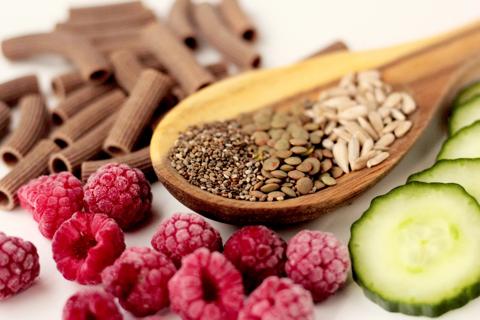 Wooden spoon full of brown rice, chia seeds and lentils, with pasta, cucumber slices and raspberries on table