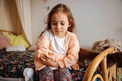 Young child sitting on edge of bed, holding stomach in discomfort
