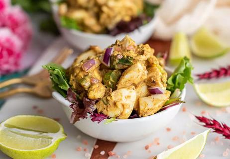 A small bowl holds curry egg salad with eggs, onions and lettuce on a cutting board beside sliced limes.