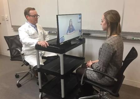 A patient looks at a woman in a monitor while a doctor in a white lab coat watches.