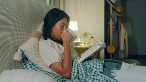 Person drinking from a mug while in bed reading a book