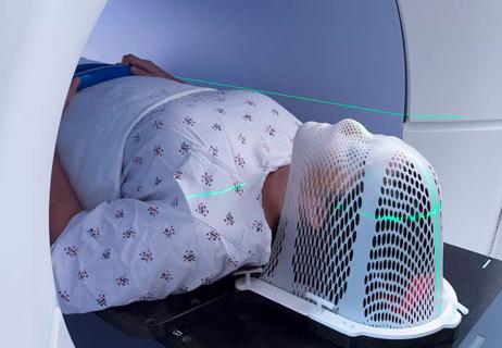 Person lies in radiation machine with face covered by protective netting as lasers scan body