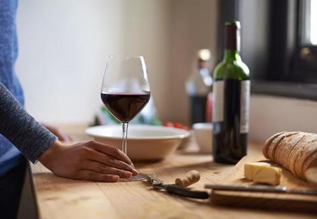 Hand holding a glass of wine atop a wooden counter, with wine and bread in background