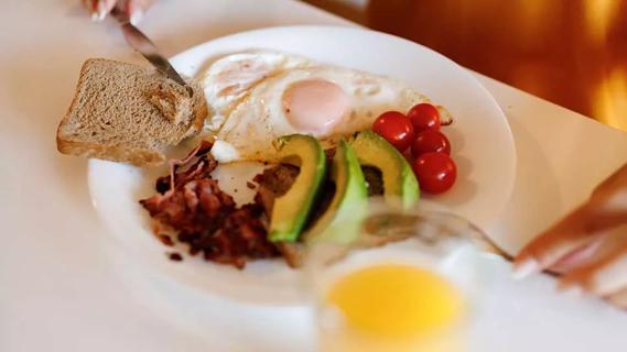 close-up of plate with eggs, wheat toast, avocados, hands holding knife and fork
