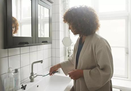 A person in a bathrobe running water over their toothbrush in a sink