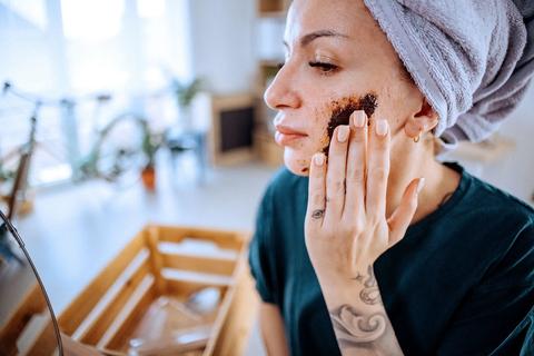 Person with wet hair wrapped in towel, applying exfoliating scrub to their face