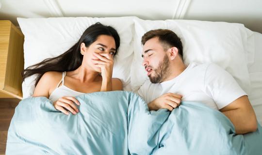 Couple in bed, with one covering their mouth as the other sleeps with their mouth open