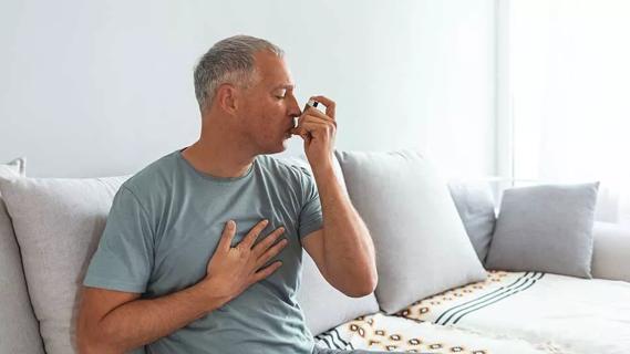 male sitting on couch using inhaler and holding chest