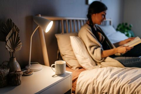 Person sitting in bed in the evening, reading a book, with cup of tea on bedside table